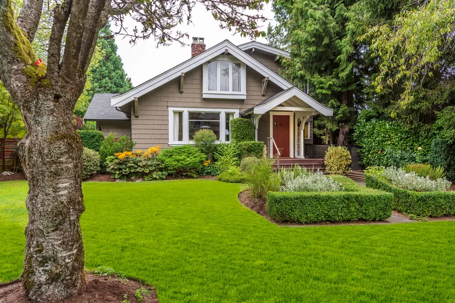 A single-story Craftsman bungalow with taupe shingle siding, white trim and a red front door, set behind clipped hedges and a wide mown lawn
