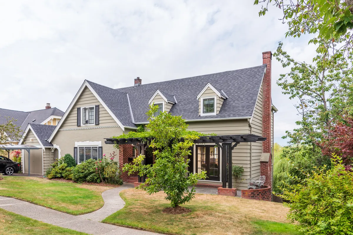 A one-and-a-half-story cottage with sage lap siding, two roof dormers and a brick pergola over the entry, reached by a curved concrete path