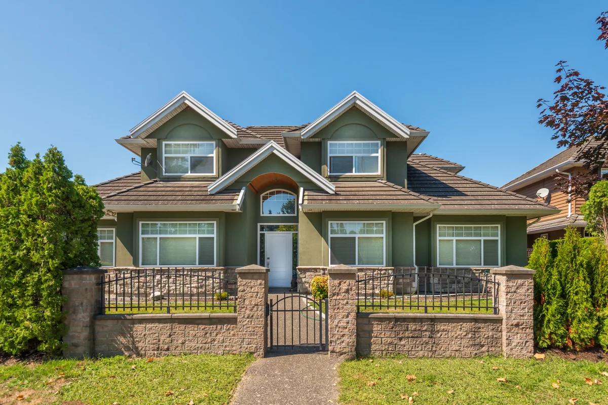 A two-story house with sage-green stucco, twin front gables and a tile roof, standing behind a low block wall with an iron gate