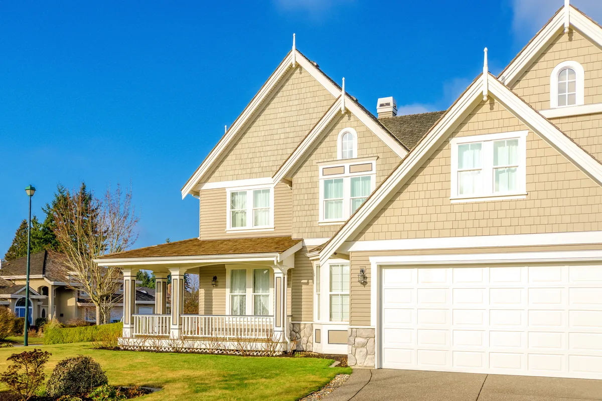 A two-story house with tan shingle siding, steep gables, a covered front porch and an attached two-car garage
