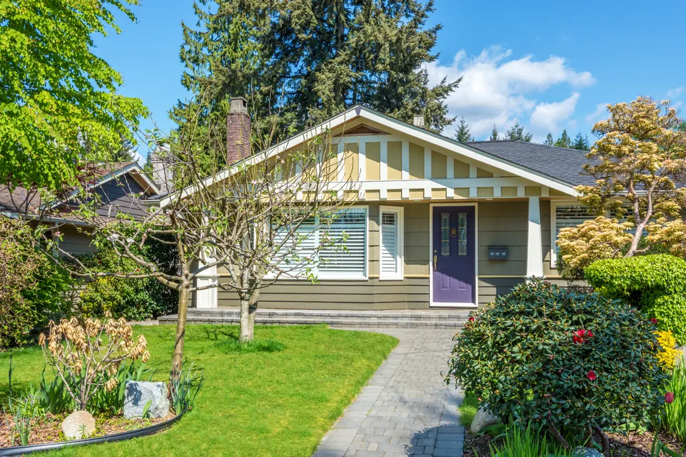 A single-story bungalow with olive and cream lap siding, a wide front gable and a panelled front door, reached by a paver walkway through planted beds