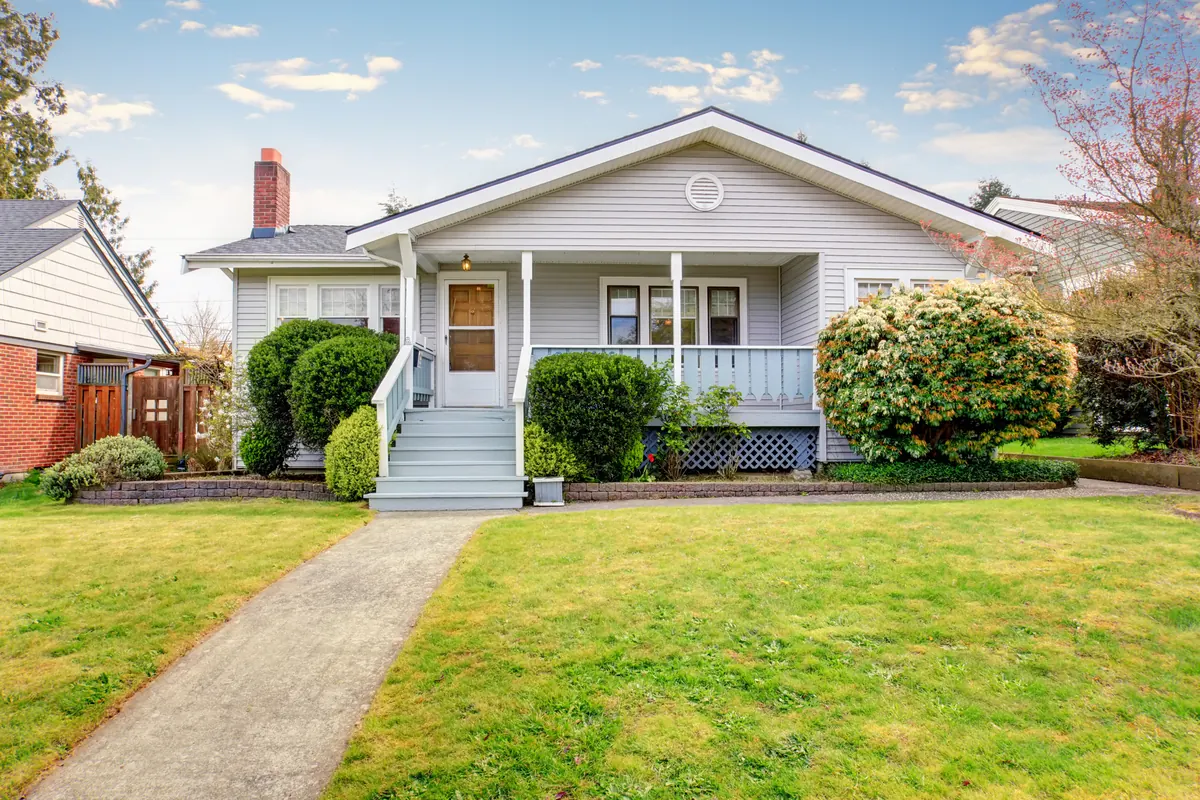 A single-story gray bungalow with a wide front gable and a covered porch with pale blue railing, set back behind a concrete walkway