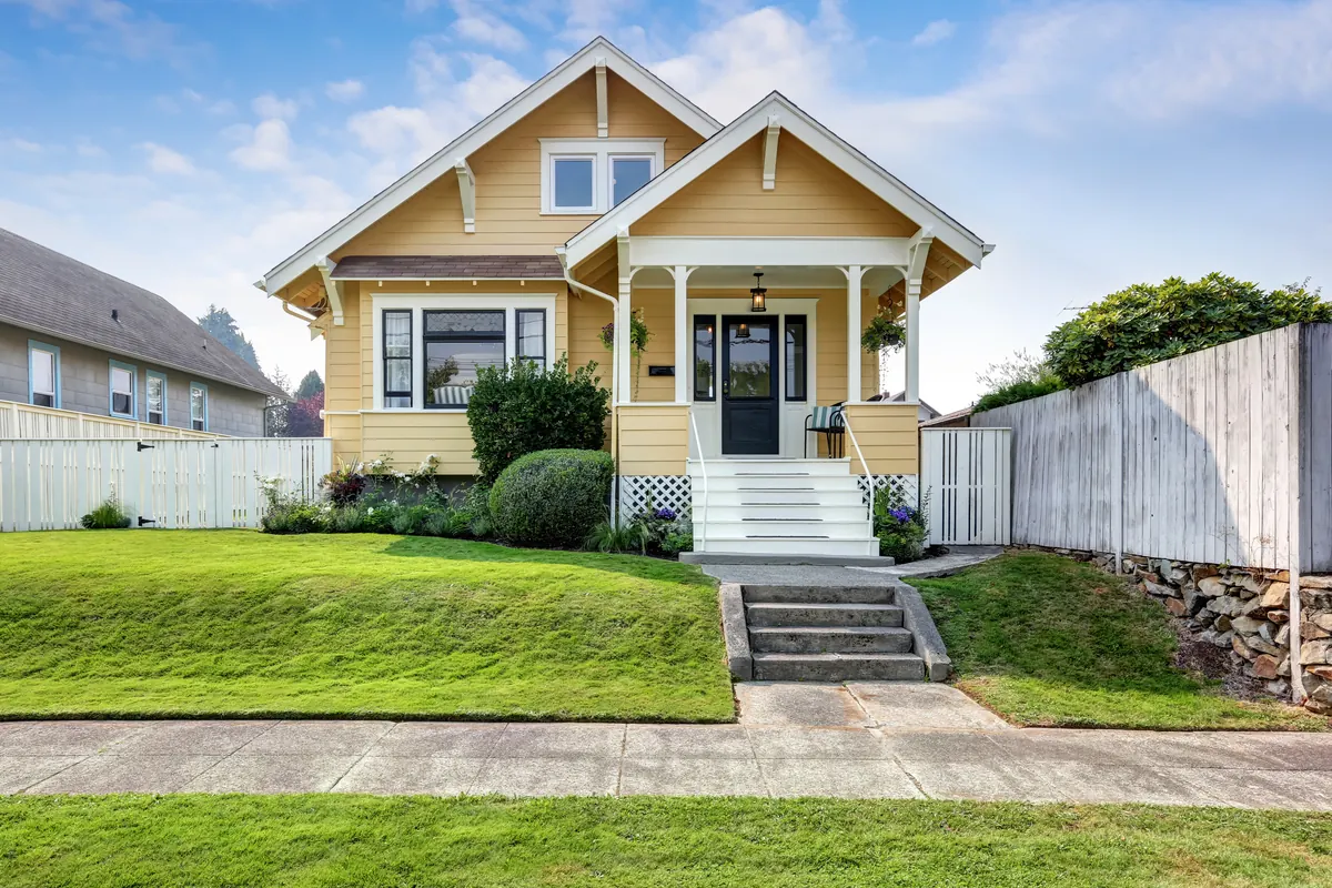 A yellow Craftsman bungalow with white trim, a gabled entry porch on square columns and a bay window, above a sloped lawn reached by concrete steps